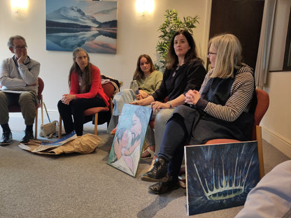 Group of people sitting in a circle with art materials and paintings in a room.